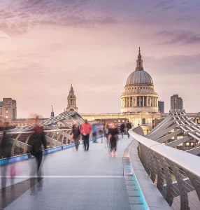 A busy bridge in London with a purple sunset in the sky and blurred people crossing the bridge.